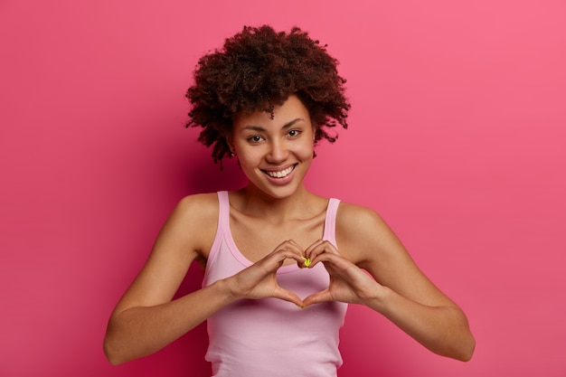 Flirty happy young curly woman expresses love and affection, makes heart gesture, shows what you mean to her, thanks dear friend for help, has charming tender look, poses against rosy wall