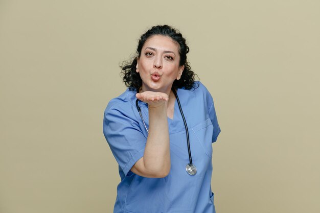 Flirty middleaged female doctor wearing uniform and stethoscope around neck keeping hand behind back looking at camera blowing kiss isolated on olive background
