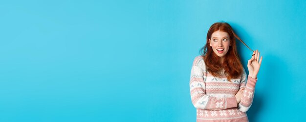 Flirty redhead girl staring left playing with hair strand and thinking standing over blue background