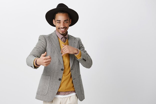 Flirty romantic male wanting have fun. satisfied confident african-american man in stylish black hat and jacket, making sensual and passionate gestures, smiling curiously over gray wall