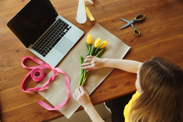 Florist at work: woman shows how to make bouquet with tulips. Young caucasian woman gives online workshop of doing gift, present for celebration. Working at home while isolated, quarantined concept.