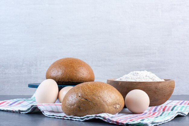 Flour bowl, eggs and buns on a towel on marble.