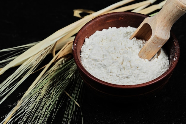 Flour in bowl with wheat on dark table, close up.