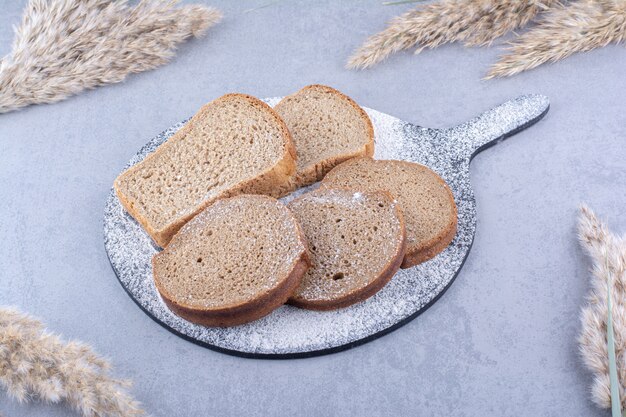 Покрытая мукой доска с ломтиками хлеба рядом со стеблями ковыля на мраморной поверхности