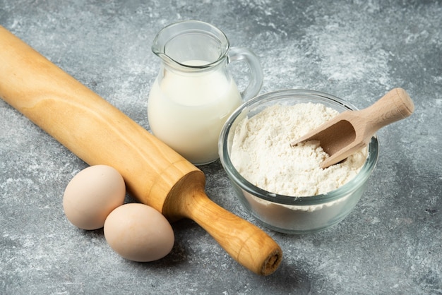 Flour, eggs, milk and rolling pin on marble surface.