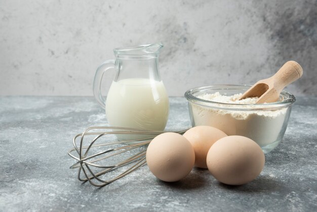 Flour, eggs, milk and whisker on marble table.