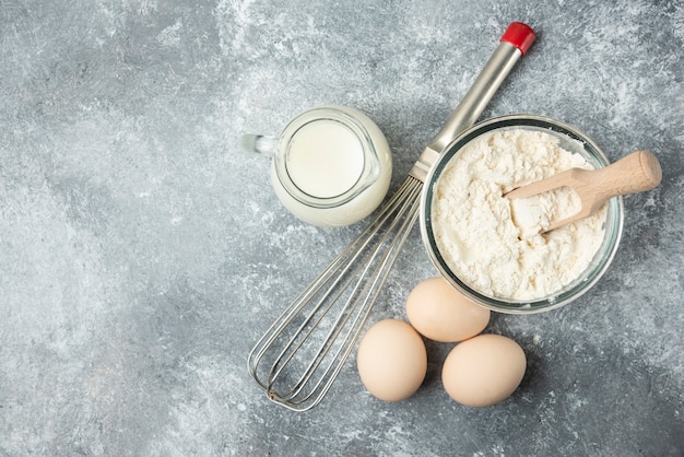 Flour, eggs, milk and whisker on marble.