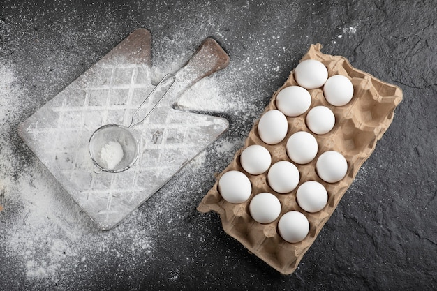 Flour spilled on wooden board and raw eggs on black surface