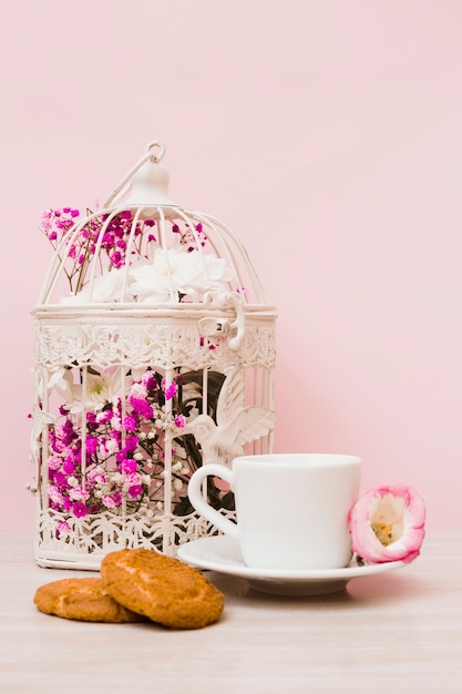 Flower cage; coffee cup and cookies on wooden desk against pastel pink background