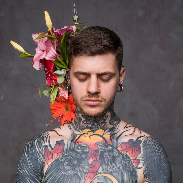 회색 배경으로 문신을하고 피어싱 된 젊은 남자 뒤에 꽃 장식