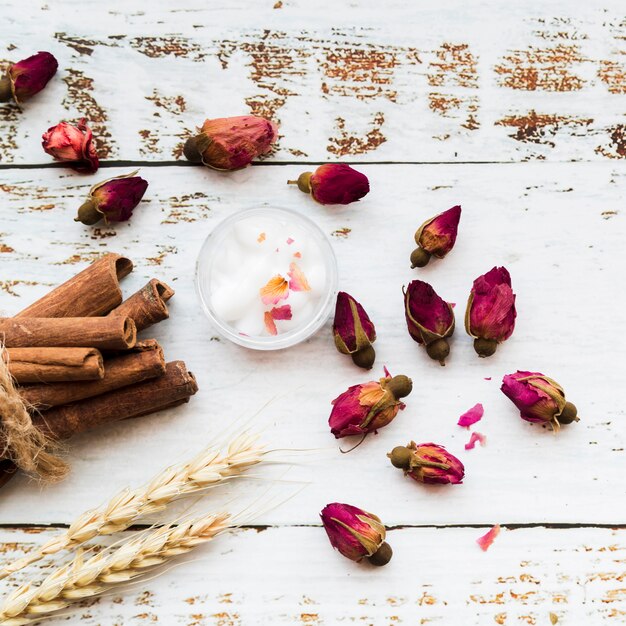 Цветочный чай из бутонов роз; палочки корицы; хлопок в миске; сноп колосьев пшеницы на белой деревянной доске