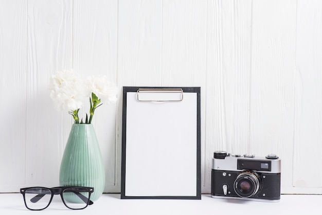 Flower vase; eyeglasses; camera and blank paper on clipboard against blank wooden wall