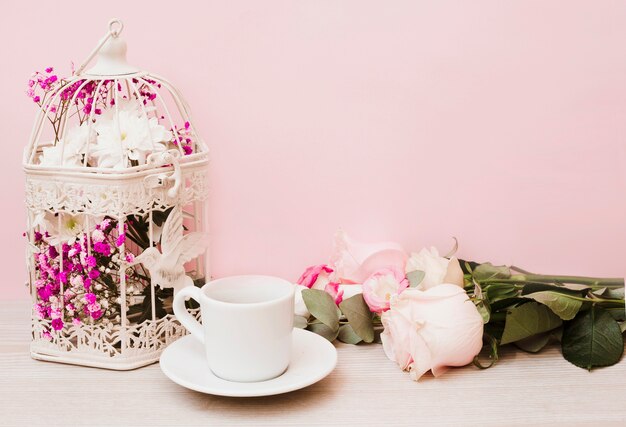 Flowers in antique cage; cup; saucer and roses on wooden table against pink backdrop