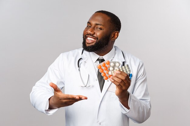 Flu, disease, healthcare and medicine concept. Happy african-american male doctor in white coat present new drugs, cure from disease or viruses, showing pills guarantee good quality of treatment