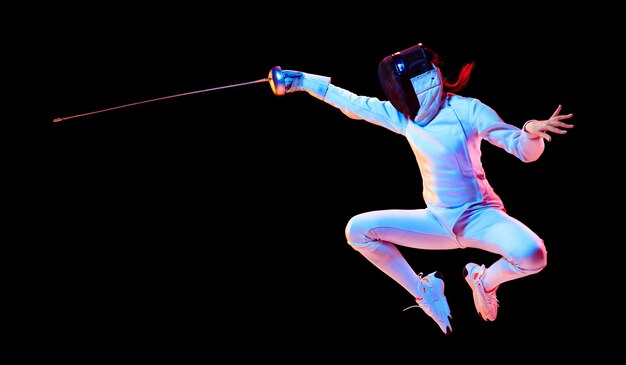 Flying. Teen girl in fencing costume with sword in hand isolated on black wall, neon light. Young model practicing and training in motion, action. Copyspace. Sport, healthy lifestyle. Flyer.