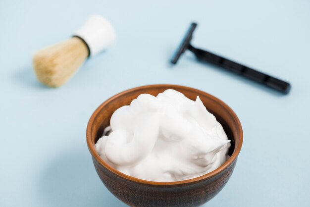 Foam in wooden bowl in front of razor and shaving brush against blue background