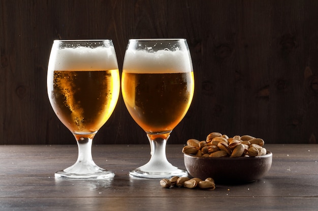 Foamed beer with pistachio in goblet glasses on wooden table, side view.