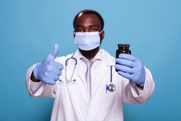 Focus on doctor holding pill bottle giving thumbs up wearing face mask and latex gloves. detail of medic doing hand gesture approving prescription antibiotics dressed in hospital uniform.