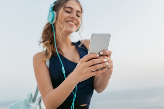 Focus on hands of happy young woman in sportswear chatting on phone, listening to music through headphones on sea. Smiling, expressing true positive emotions