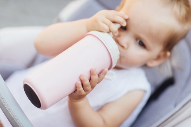 Focus at pretty girl sitting in a stroller and drinking water or milk from her thermos