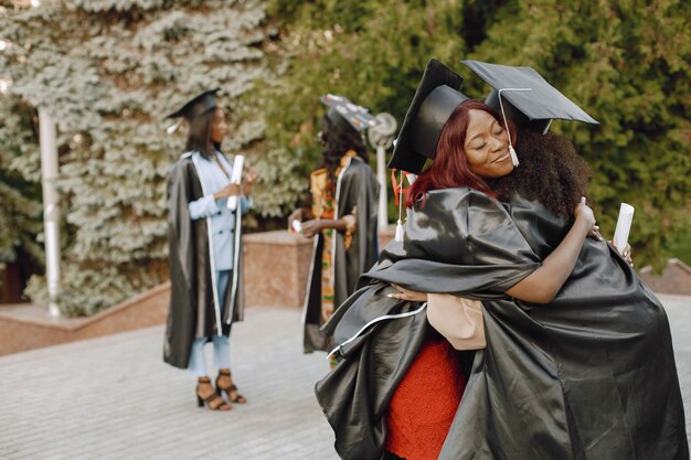 黒の卒業式のガウンに身を包んだ2人の若いアフリカ系アメリカ人の女子学生に焦点を当てます。背景としてのキャンパス。抱き締める女の子