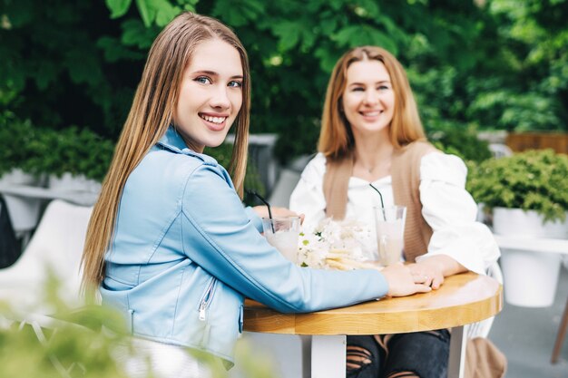 In focus, a young blonde girl is smiling at the camera and holding her mother's hand