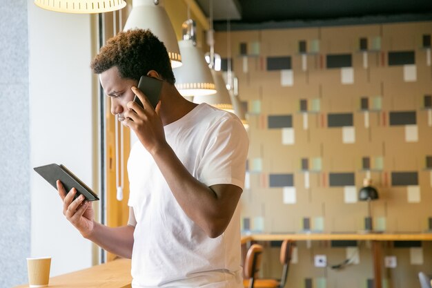 Сосредоточенный афро-американский парень разговаривает по мобильному телефону и смотрит на экран планшета