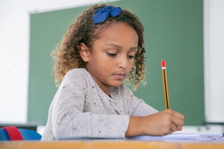 Free Photo focused african american primary school girl sitting at desk and writing in pencil