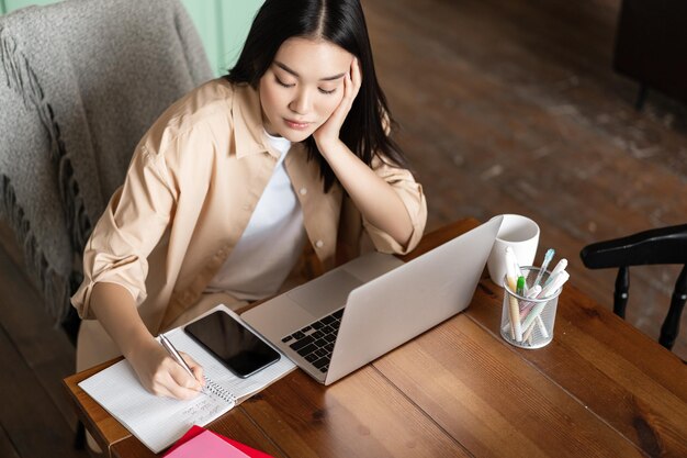 Focused asian girl doing homework writing notes and sitting near laptop attend webinar online course...