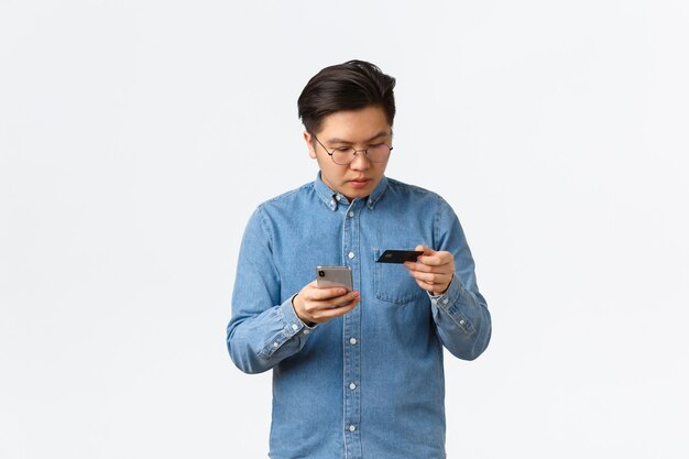 Focused asian guy in glasses typing credit card number to make online purchase, using mobile phone and bank account to buy something in internet store, standing white background.