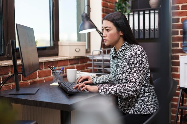 Focused asian marketing company office worker using work computer to analyze department management chart. Finance agency employee reviewing startup audit results while in modern workspace.