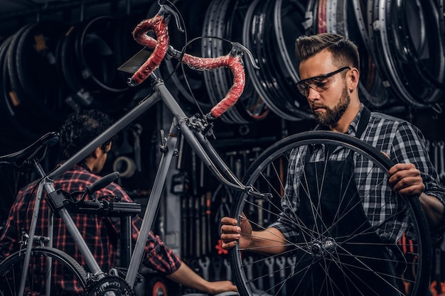 안경을 쓴 집중된 매력적인 남자가 바쁜 작업장에서 자전거 바퀴를 연결하고 있습니다.