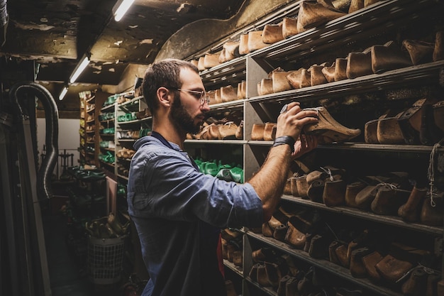 Focused bearded cobbler is looking for right shoe form at the storage.