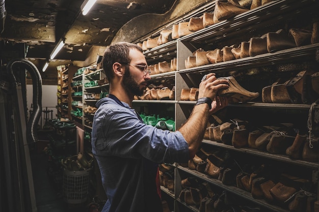 Сосредоточенный бородатый сапожник ищет на складе нужную форму обуви.