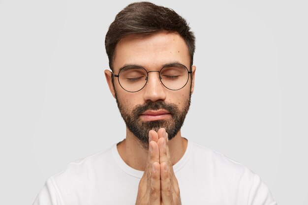 focused bearded young man model keeps palms in praying gesture, believes in good fortuney.
