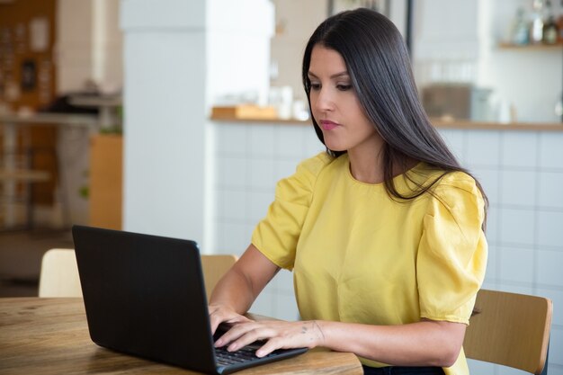 Focused beautiful black haired woman sitting at table in co-working space, using laptop, looking at display and typing