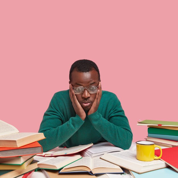 집중된 흑인 남성은 열린 책을 응시하고 뺨을 만지며 놀랍게도 학습 주제를 보며 녹색 점퍼를 입습니다.