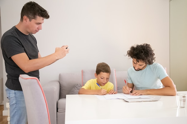 Сосредоточенный мальчик делает школьное домашнее задание с помощью двух пап, пишет в бумагах. Человек фотографирует свою семью. Концепция семьи и родителей-геев