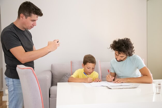 Focused boy doing school home task with help of two dads, writing in papers. Man taking picture of his family. Family and gay parents concept