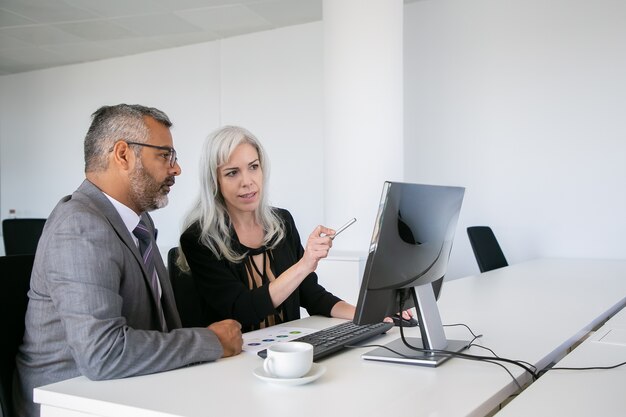 Focused business colleagues watching content on computer, pointing at display and talking while sitting at desk with paper chart. Business communication concept