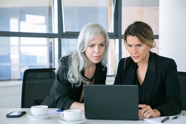 Focused business ladies looking at laptop display while sitting at table with cups of coffee in office. Teamwork and communication concept