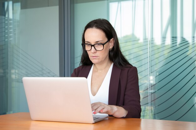 眼鏡とジャケットを着て、オフィスのコンピューターで働いて、テーブルで白いラップトップを使用して焦点を当てたビジネス女性