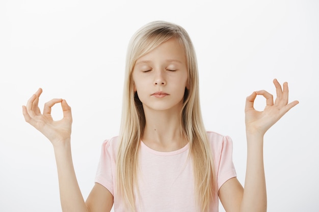 Focused calm european girl trying meditation with mom. Portrait of confident good-looking child with blond hair, closing eyes and standing over gray wall in yoga pose with zen gestures