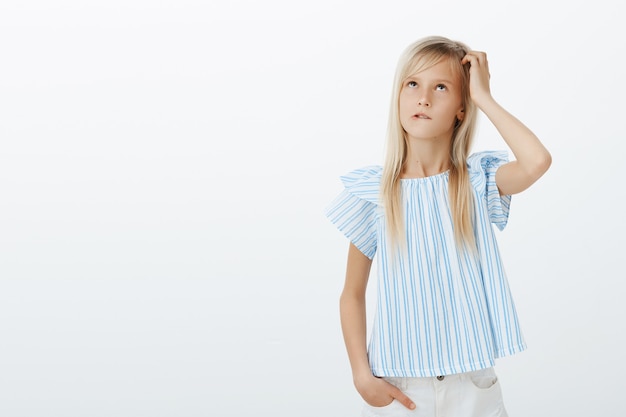 Focused clueless girl trying to calculate in mind near blackboard. Portrait of confused questioned kid in stylish blue blouse, scratching head and looking up, biting lip while thinking over gray wall