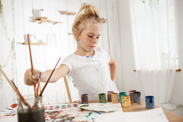 Focused on creative process cute little blond with hair bun and freckled face in white t-shirt in art room.