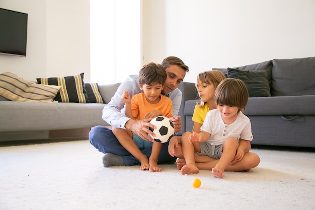 Focused dad holding ball and talking with children. Loving Caucasian father and kids sitting on carpet in living room and playing together. Childhood, game activity and fatherhood concept