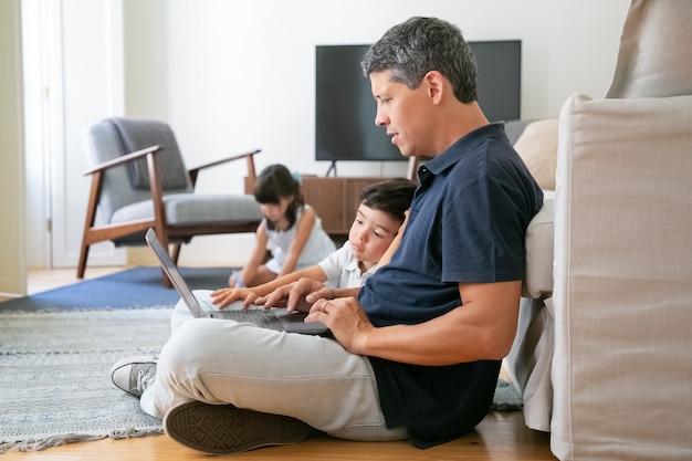 Focused dad and little son sitting on floor in apartment, using laptop, working or watching content.