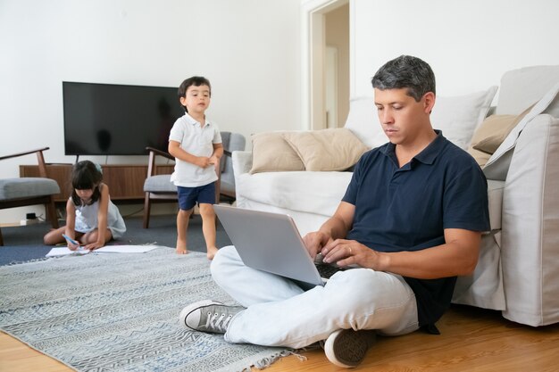 Сосредоточенный отец сидит на полу и использует компьютер, пока его маленькие дети рисуют и гуляют