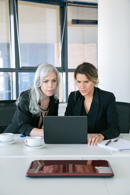 Сосредоточенные взволнованные деловые женщины, глядя на дисплей ноутбука, сидя за столом с чашками кофе в офисе. Передний план. Концепция совместной работы и коммуникации