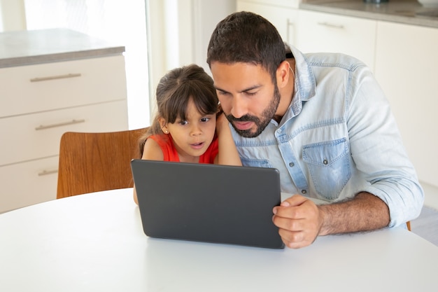 Focused excited girl and her dad using laptop, sitting at table, staring at display.